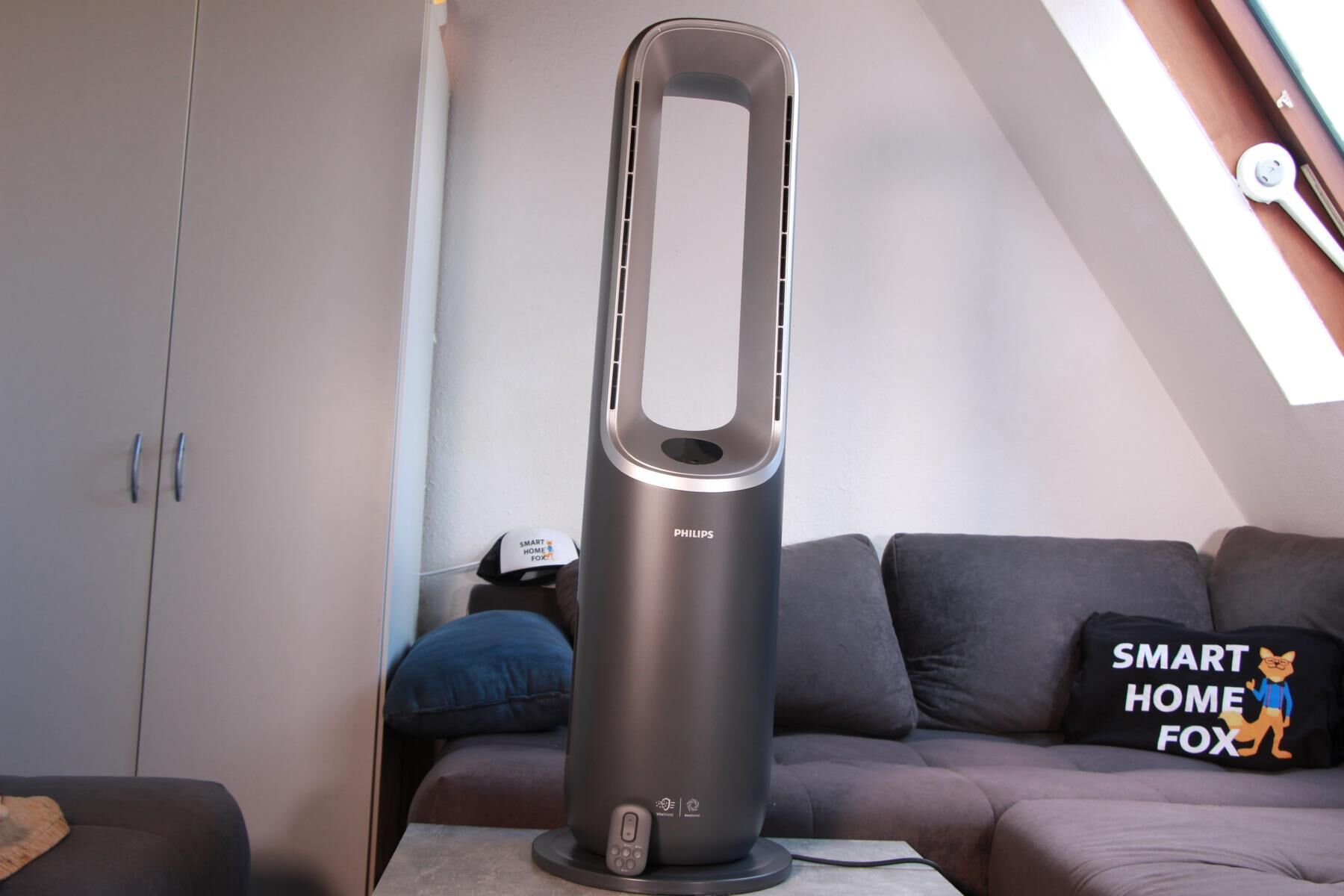 Der Philips Air Performer Series 8000 steht auf einem Couchtisch vor einer Couch und die Fernbedienung ist an den Sockel angelehnt. 